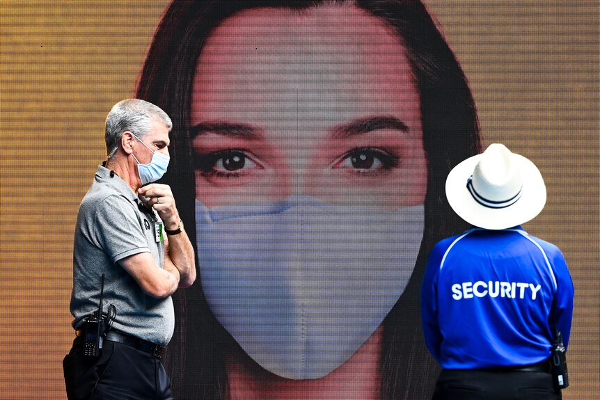 A man with security on his shirt and another with a walkie talkie stand in front of a sign with a woman with a mask