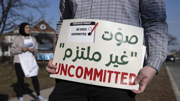 An Arab American voter in Michigan stands holding a sign reading 'Uncommitted' with accompanying Arabic script
