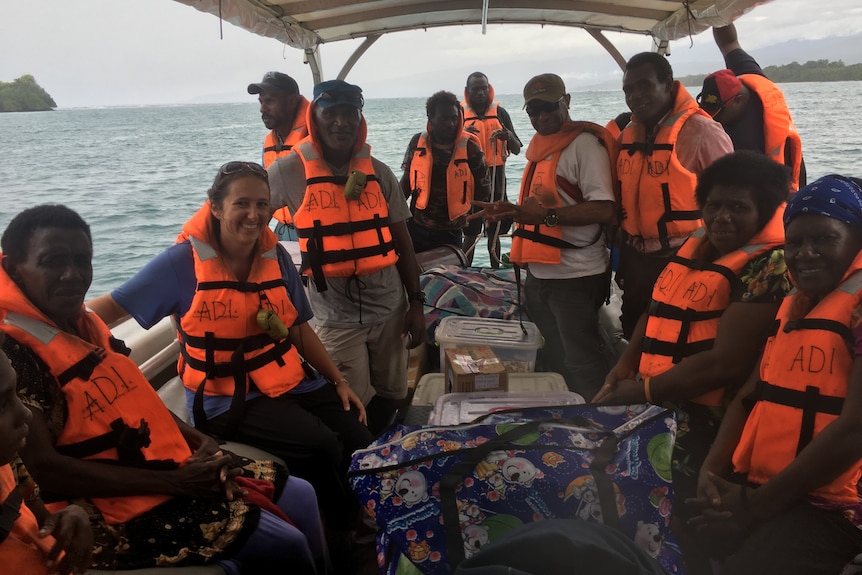 Several men and women smile at camera wearing bright orange life vests aboard boat that is in the water.