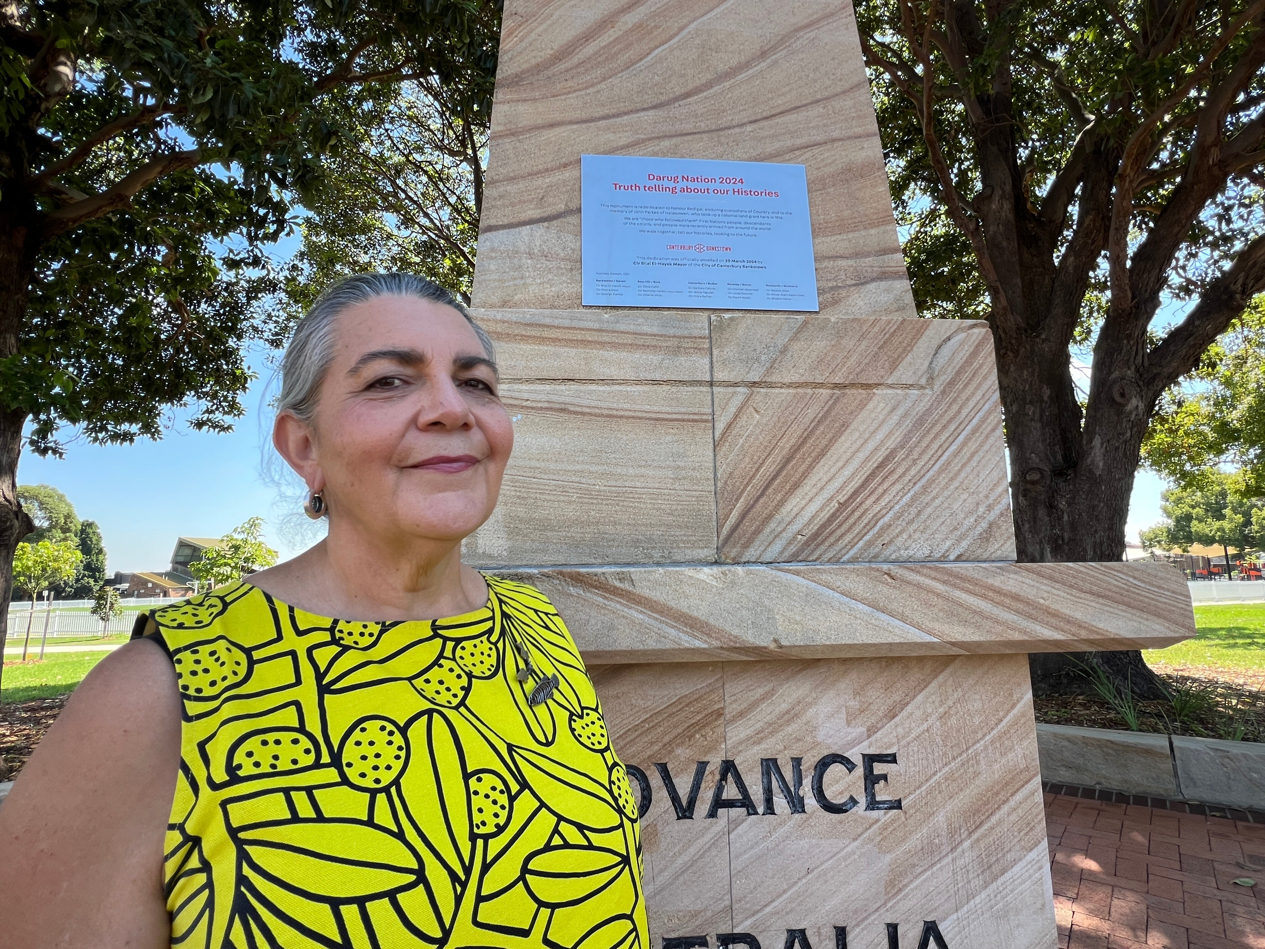 A photo of Jennifer Newman in front of the monument plaque. She has greying hair tied in a bun. She's wearing a yellow top.