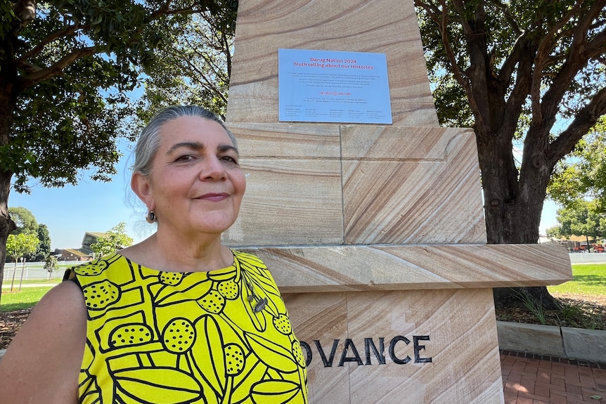 A photo of Jennifer Newman in front of the monument plaque. She has greying hair tied in a bun. She's wearing a yellow top.