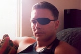 Parcel bomb victim Brett Boyd, wearing an eye patch and a black singlet, sits on a couch in his home.