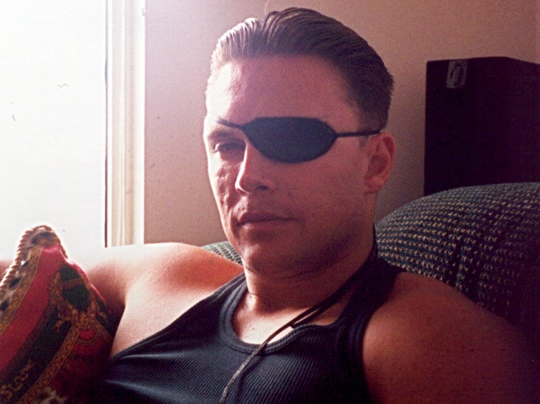 Parcel bomb victim Brett Boyd, wearing an eye patch and a black singlet, sits on a couch in his home.