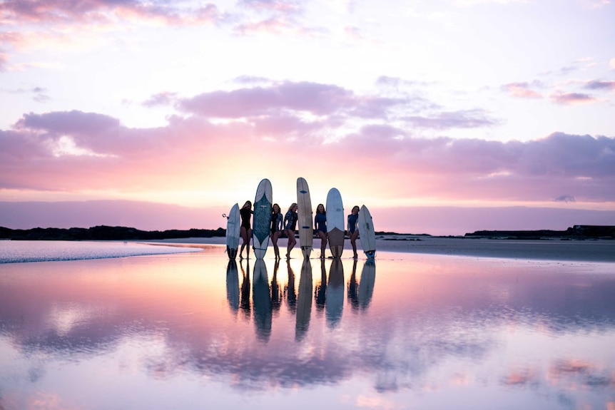 five female surfers stand on the beach holding their boards