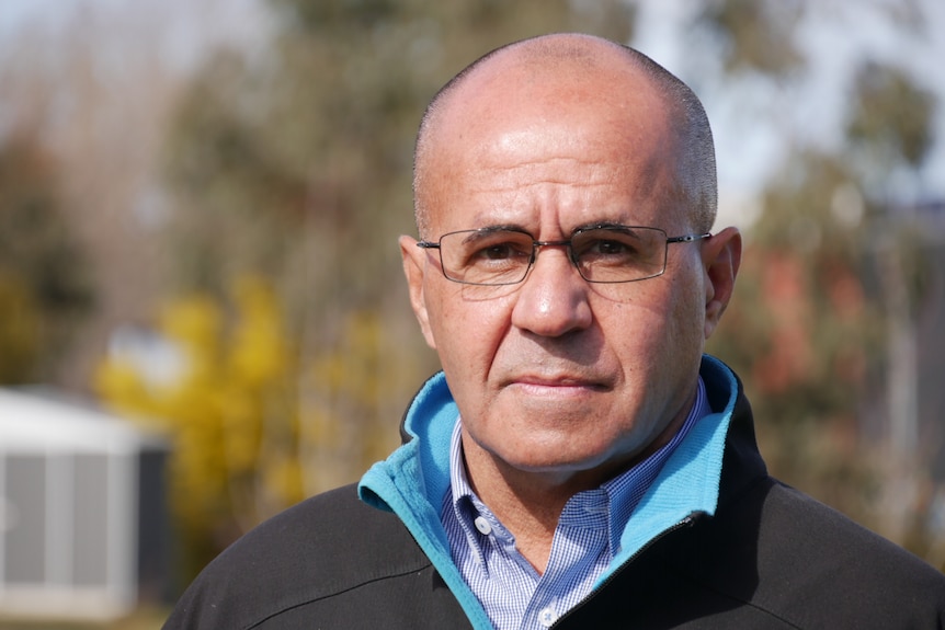 Bald man wearing dark grey jacket and glasses, standing outside.