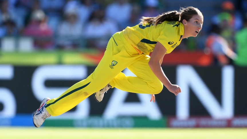 A woman in yellow follows through after bowling a cricket ball.