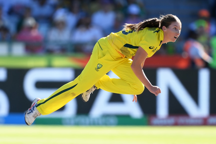 A woman in yellow follows through after bowling a cricket ball.