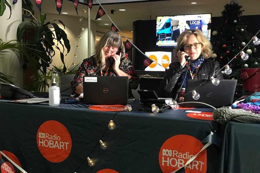 Photograph of two blong women on the phone in front of laptops, under a tent which is covered with ABC Hobart logos.