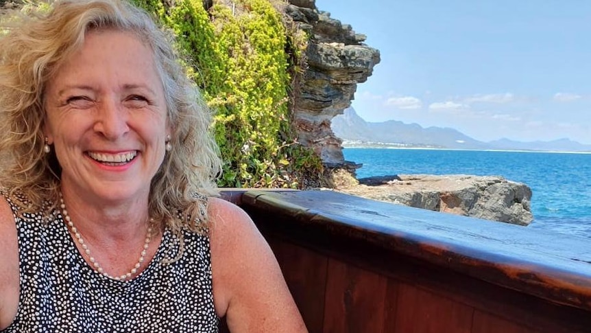 A woman with blond hair smiling with a glass of wine and a view of the sea in the background.