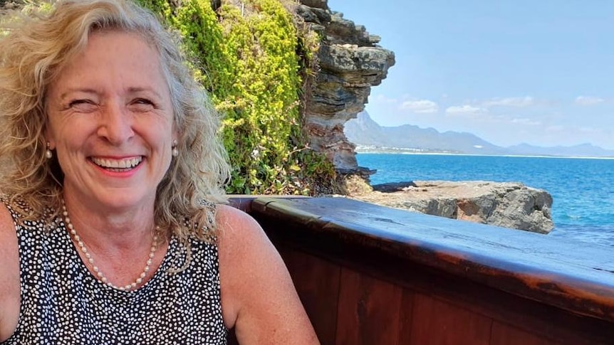 Donna dai capelli biondi sorridente con un bicchiere di vino e vista mare sullo sfondo.