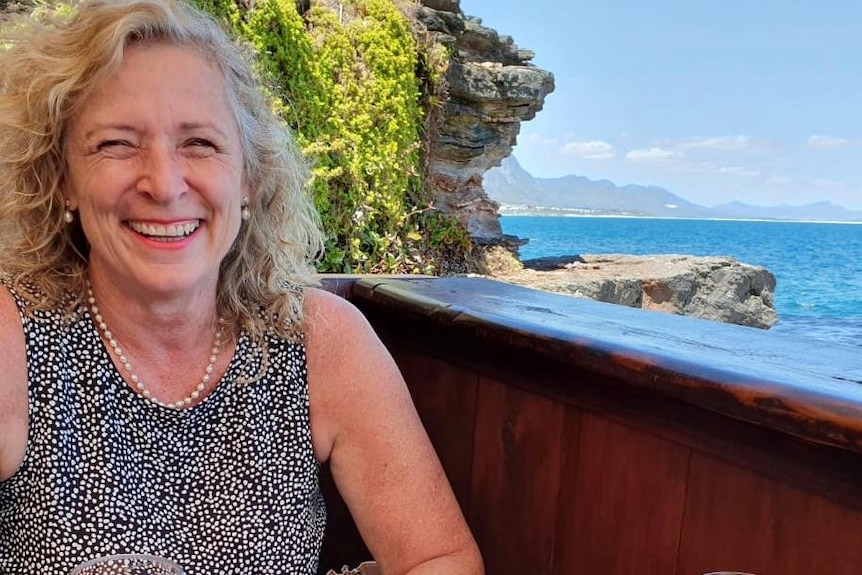 A woman with blond hair smiling with a glass of wine and a view of the sea in the background.