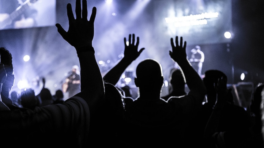 People with raised hands above their heads cast a shadow against dusty blue lights on a stage in the background