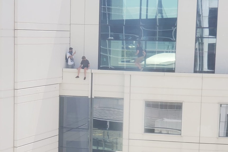 A boy takes a photo of another boy sitting on the edge of a building
