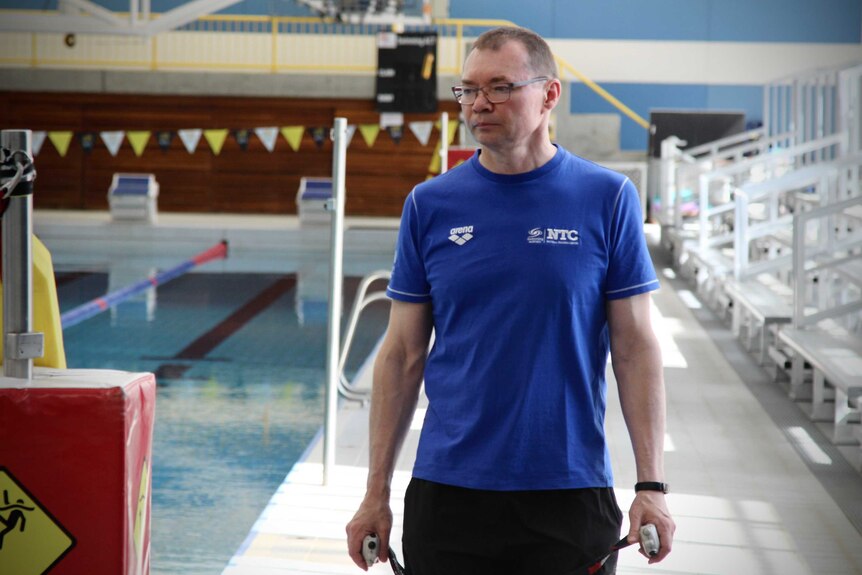 A man wearing a blue top walks alongside a pool holding a stop watch in his hand
