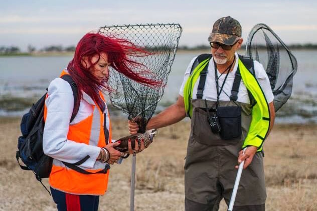 A man dressed in waders with nets and a woman holding a duck near a body of water.