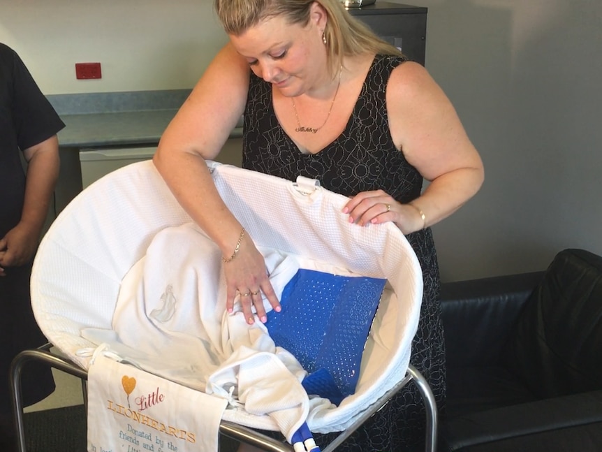 Selena Wool, standing behind a bassinet, pulls back the sheets.