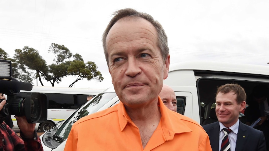 Close up shot of Bill Shorten dressed in hi-vis orange with a car, camera and two suited men behind him.