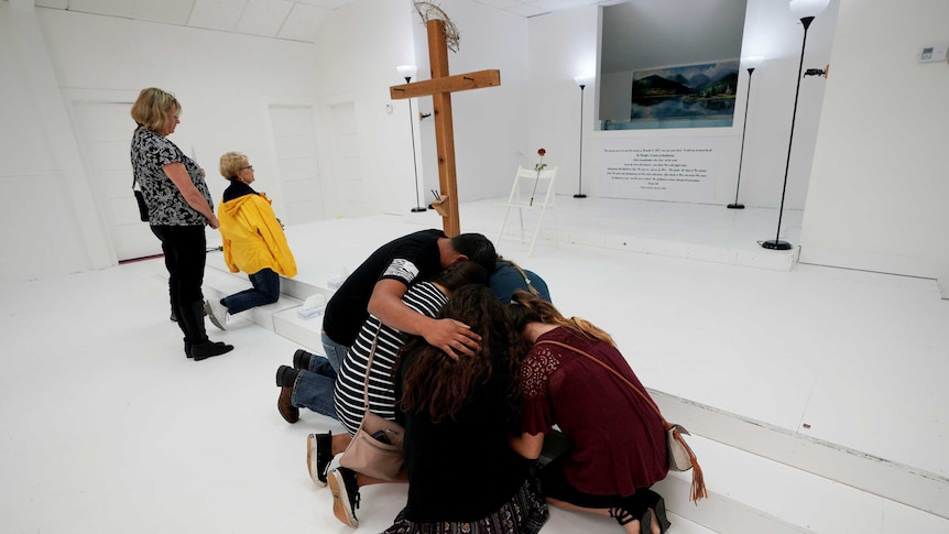 A group of people huddle and pray in front of a wooden cross as others nearby kneel and pray.