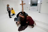 A group of people huddle and pray in front of a wooden cross as others nearby kneel and pray.