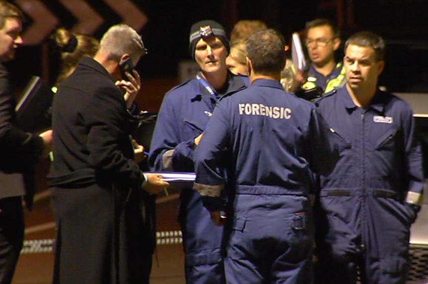 Forensic investigators in blue overalls and detectives on their phones stand in the street outside the crime scene.