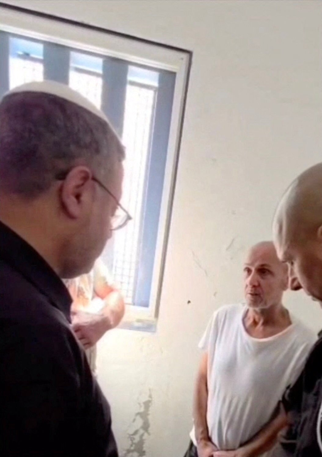 A tall Israeli man with glasses is seen looking down on a gaunt Palestinian prisoner, in handcuffs and with grey hair.