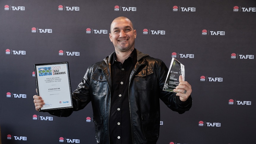 Steve Bastian wears a leather jacket and holds up a trophy in one hand and a framed certificate in the other