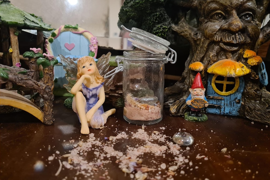 A small statue of a fairy next to a glass jar full of DIY pink fairy dust which is also spread out on the table infront