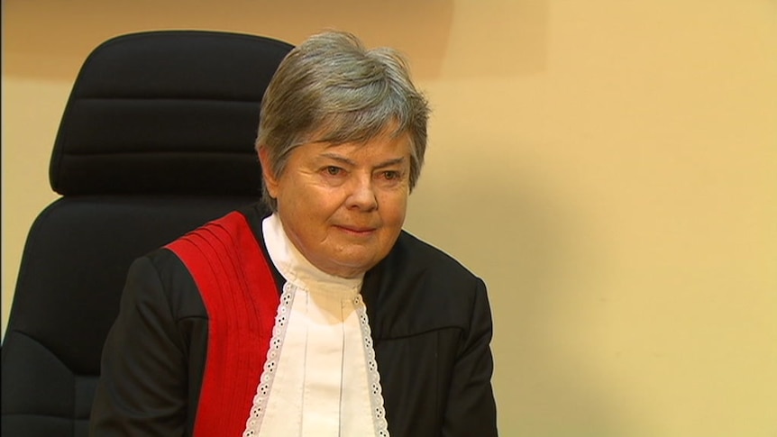 A woman sits in a chair. She is wearing a black robe with a red sash and a white collar.