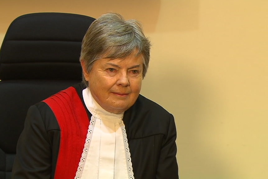 A woman sits in a chair. She is wearing a black robe with a red sash and a white collar.