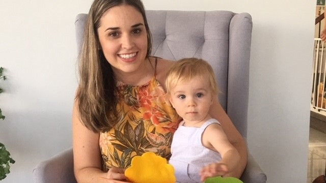 New mum Amanda Hall sits in a chair with her baby Flynn in her lap with a children's book.