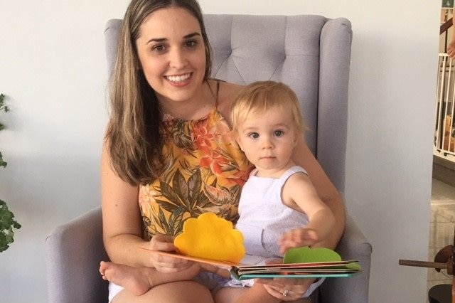 New mum Amanda Hall sits in a chair with her baby Flynn in her lap with a children's book.