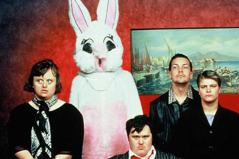 Four people look serious while standing next to someone in a rabbit costume in a burgundy coloured room.