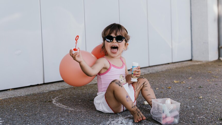 A toddler sits on the ground, smiling and holding a bubble wand 