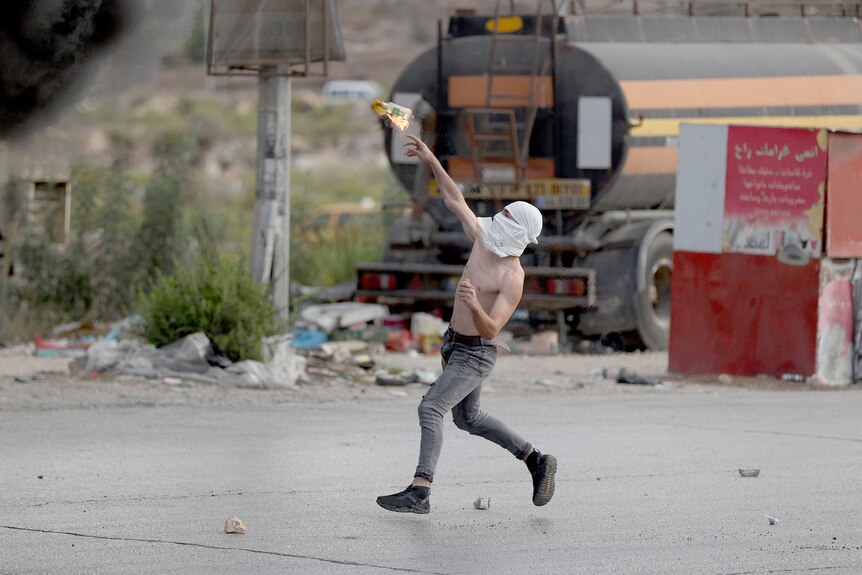 A shirtless man with a white cloth tied around his face throws a burning bottle.