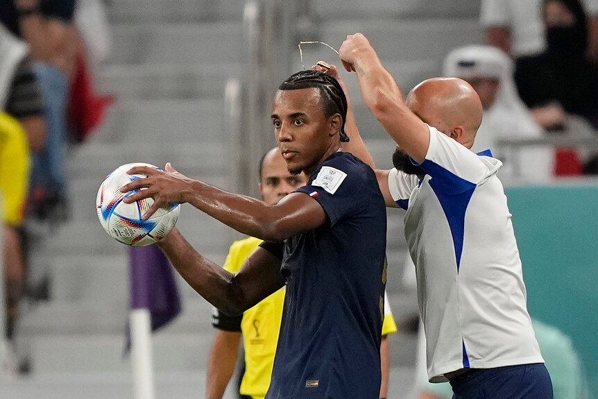 France's Jules Koundé has a necklace taken off from around his neck by an assistant coach while holding a ball at the World Cup.
