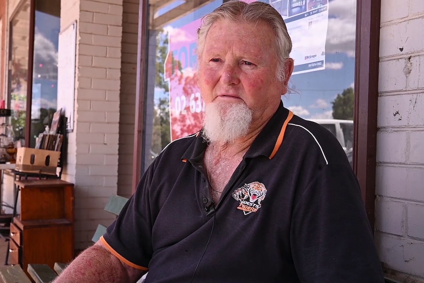 A man sits on a bench in a country town. He is wearing a Wests Tigers polo shirt.