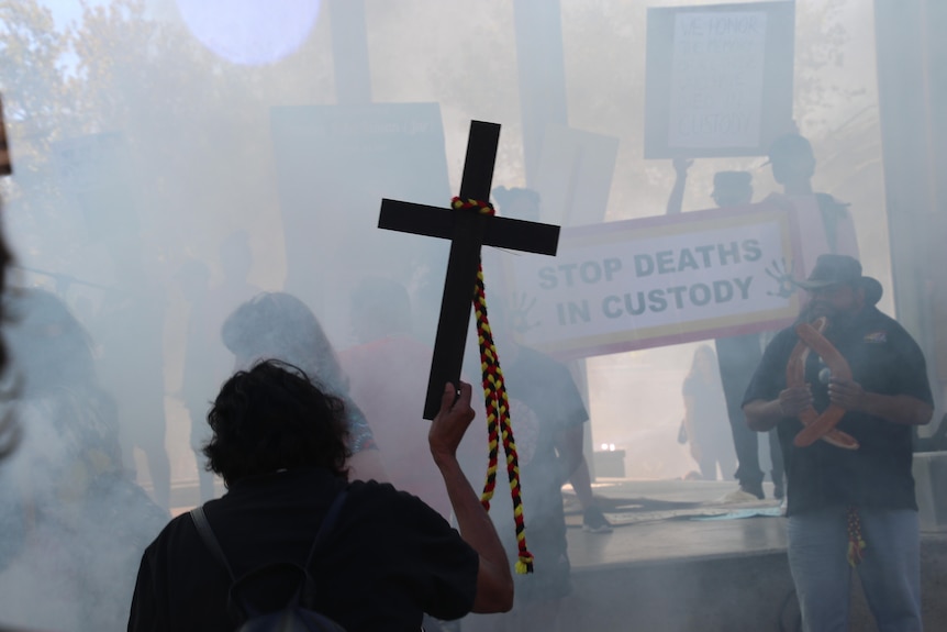 A silhouette of a woman holding a large wooden religious cross, with smoke and other people in the background. 