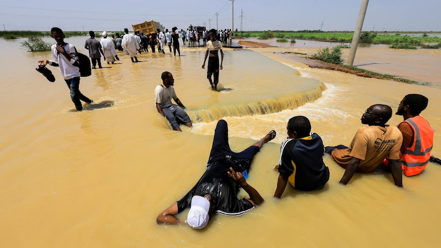 A man reclines in flood waters as others cross, carrying furniture