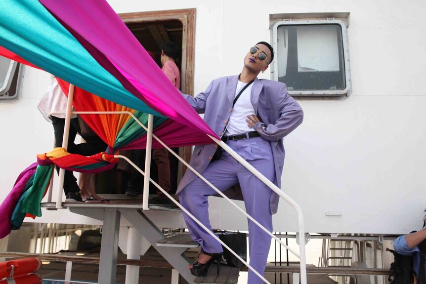 A man in a vibrant lilac suit strikes pose under a rainbow banner