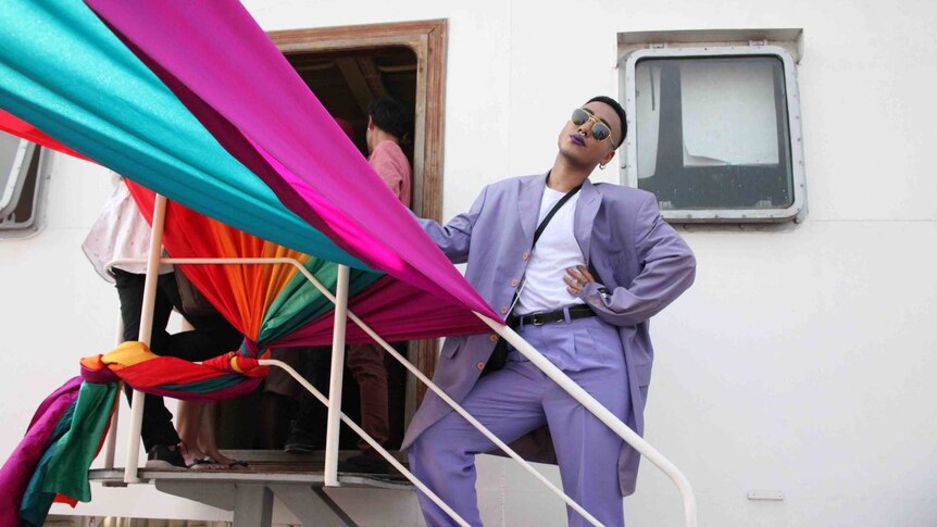 A man in a vibrant lilac suit strikes pose under a rainbow banner