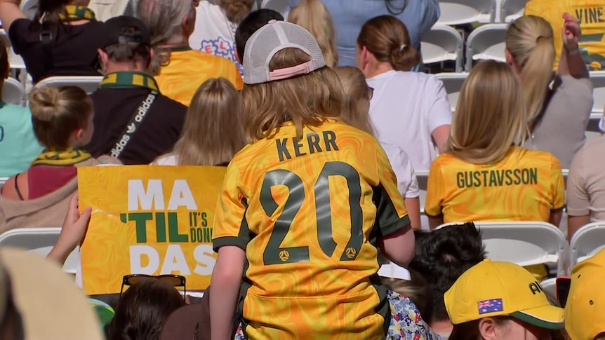 Child in the crowd wears green and gold Matildas jersey that reads 'Kerr'
