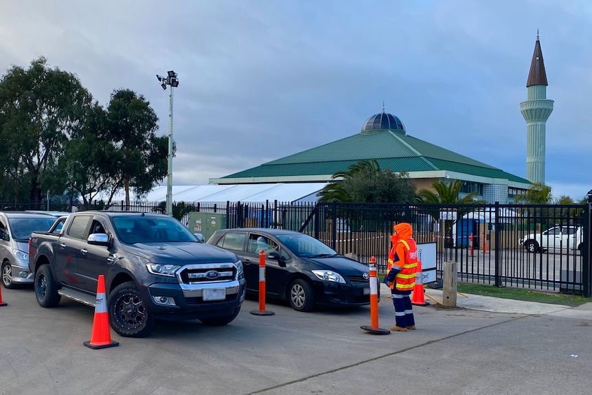 Two lines of cars queue inside witches hats outside the Al-Taqwa College campus on a cloudy morning.