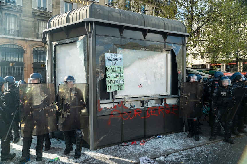 Police in rot gear stand in a line around a badly damaged bus stop