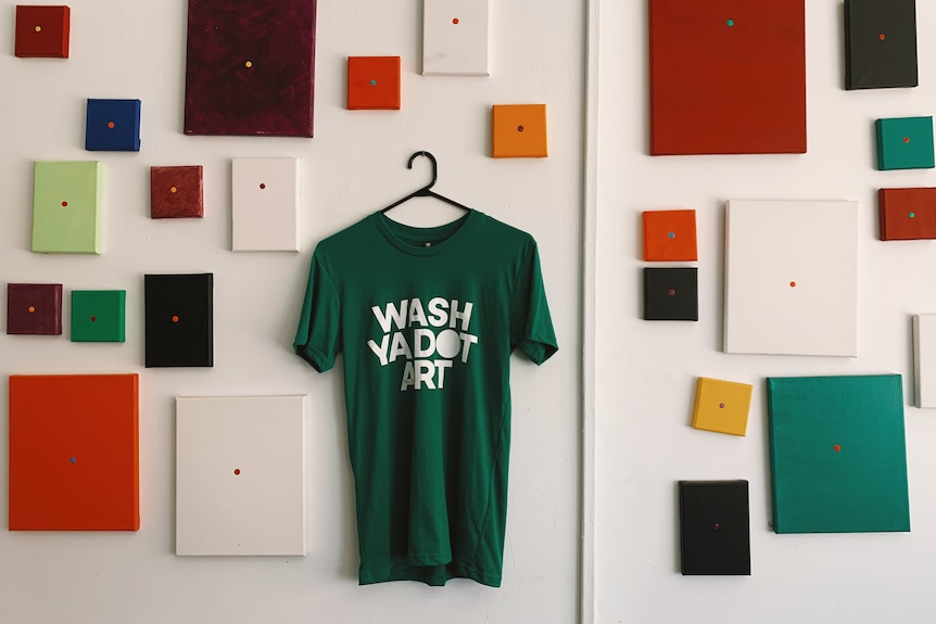Paintings hang on a wall next to a green t shirt which reads 'wash ya dot art'