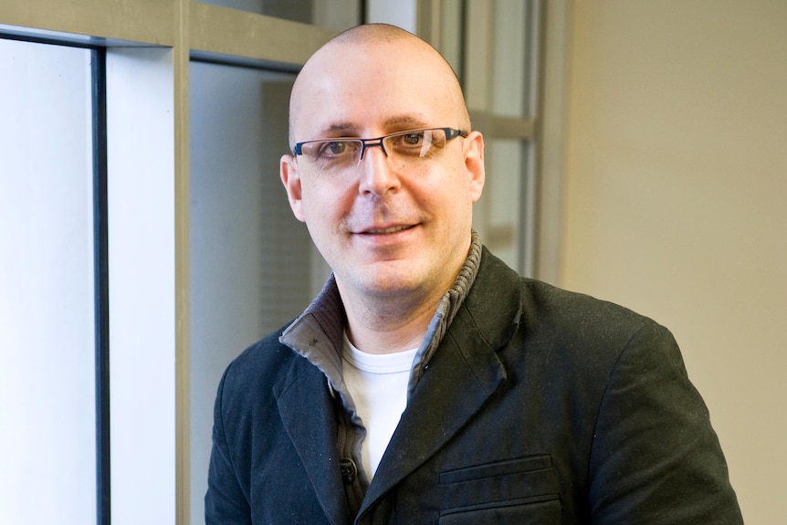 A man stands in a hallway wearing glasses.