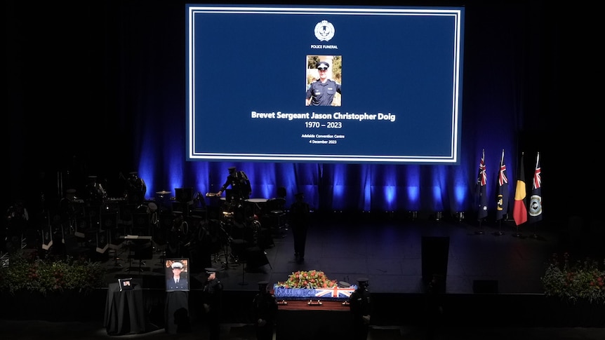 A screen with a photo of Brevet Sergeant Jason Doig, on a stage with blue lights and a coffin.