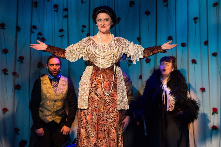 Women dressed as Dame Nellie Melba in 1920s gown with pearls and black up-do, with arms stretched as if taking a bow.