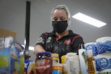 A woman wearing a mask packs various canned goods into a bag