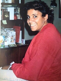 A woman wearing a red long-sleeved top looks over her shoulder at the camera smiling.
