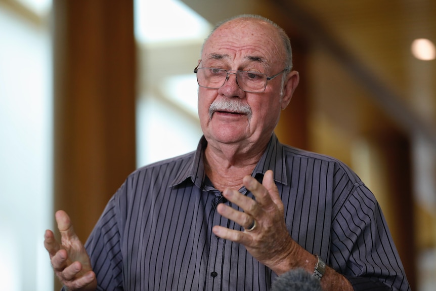 An elderly man with a large mustache holds out both arms as he speaks inward.